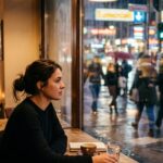 Junge Frau sitzt allein an einem Café-Fenster und blickt abends auf eine belebte Straße